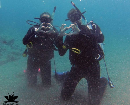 Two scuba diver doing the heart sign with the fingers while resting on their knees underwater
