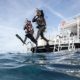 Two scuba diver exiting a boat with a gian step while holding mask and regulator with on hand and protecting the back of the head with the other hand.