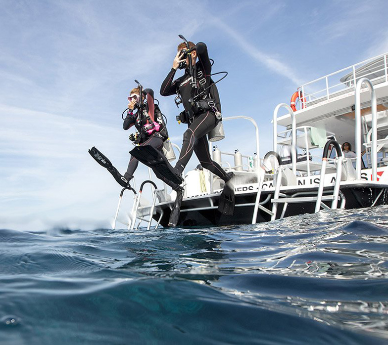 Two scuba diver exiting a boat with a gian step while holding mask and regulator with on hand and protecting the back of the head with the other hand.